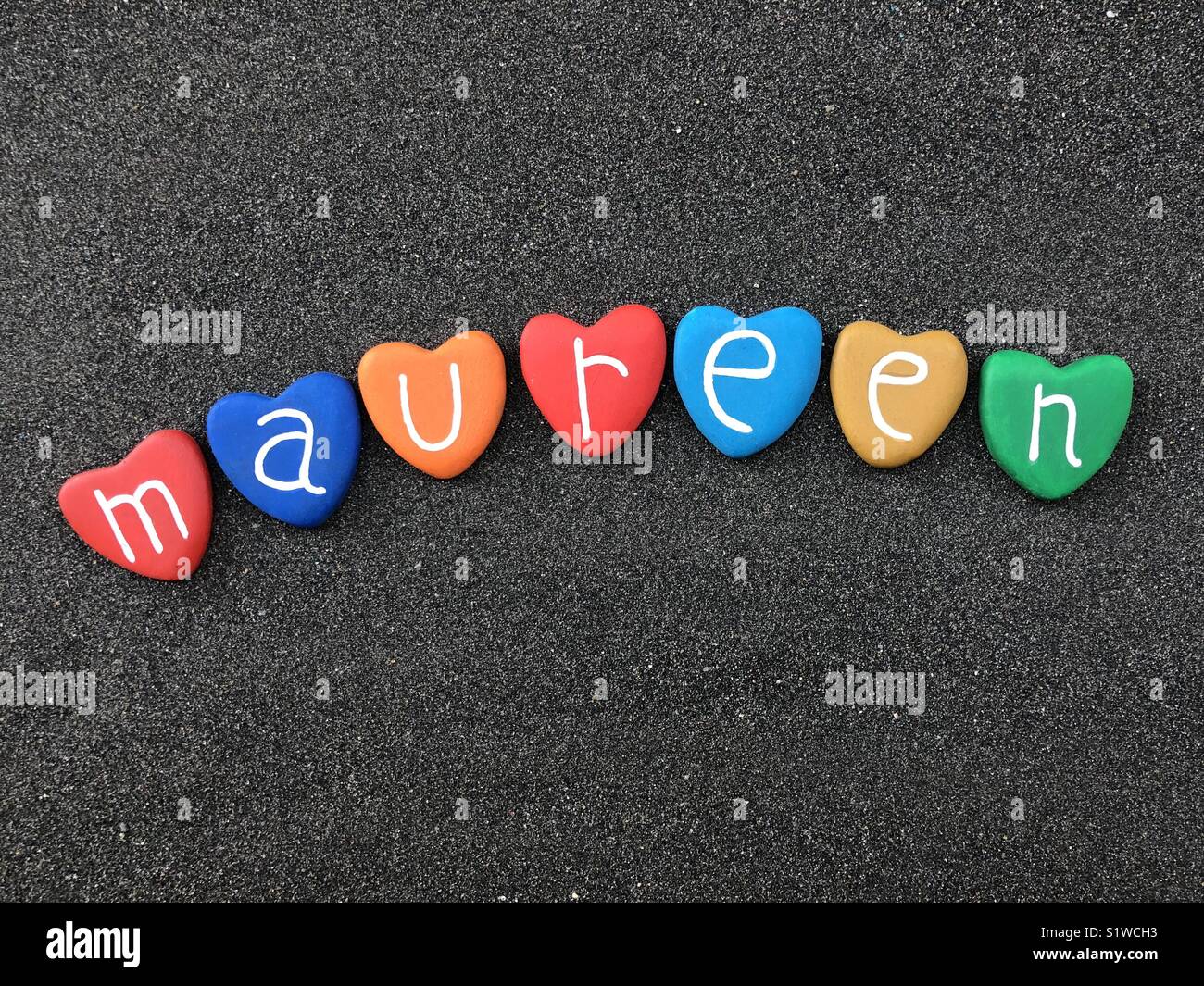 Maureen, feminine name with multicolored heart stones over black volcanic sand Stock Photo