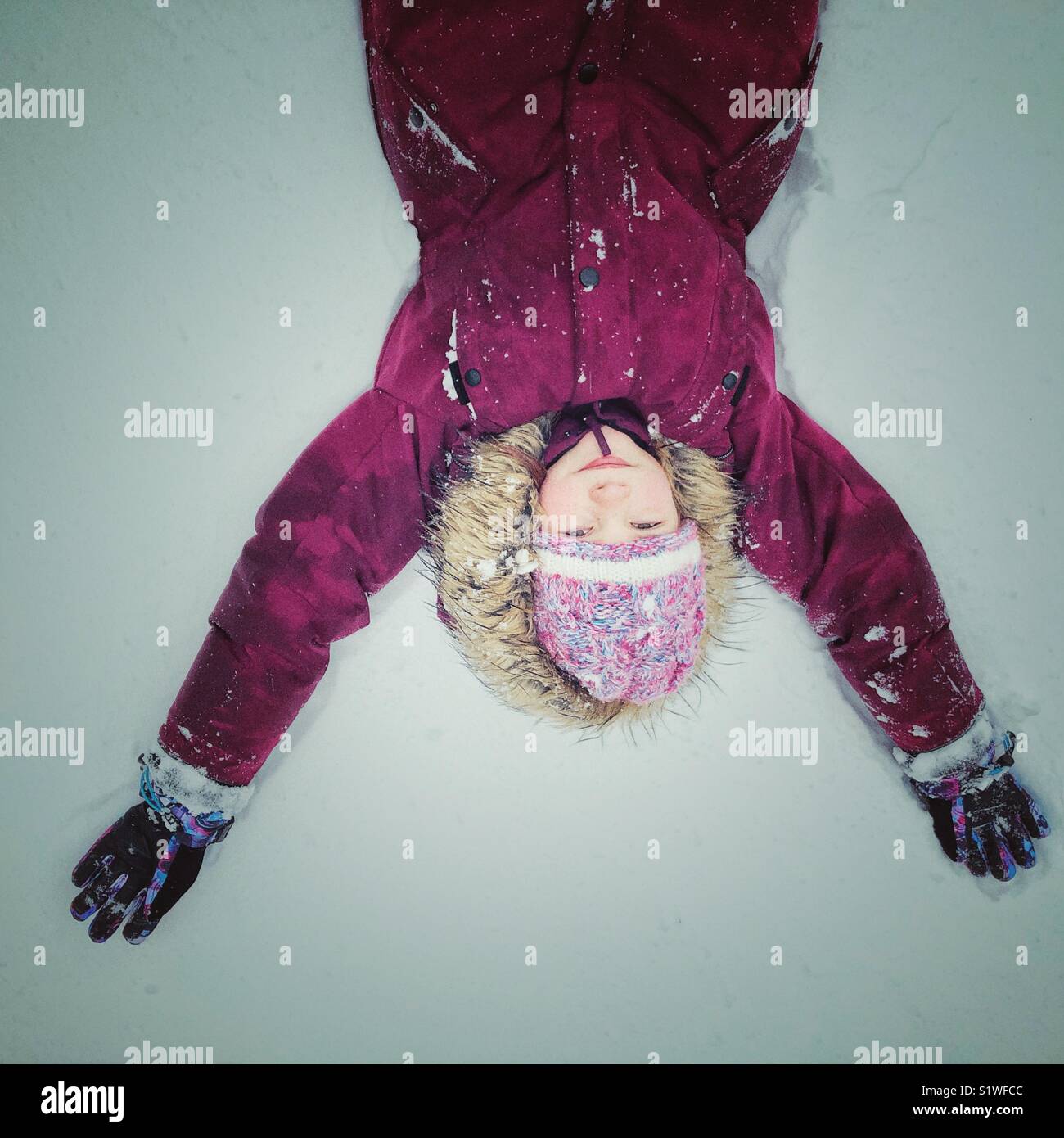 Girl lying down in snow wearing a lavender purple snowsuit looking upside down Stock Photo