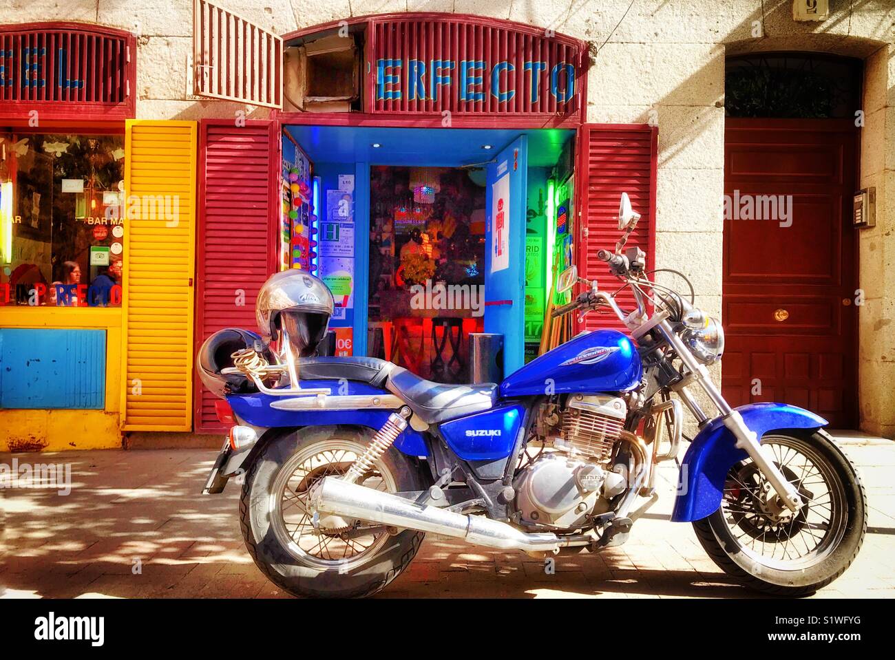 A blue and chrome Japanese Suzuki motorbike parked on the pavement in front of a colourful shop entrance in Madrid, Spain Stock Photo