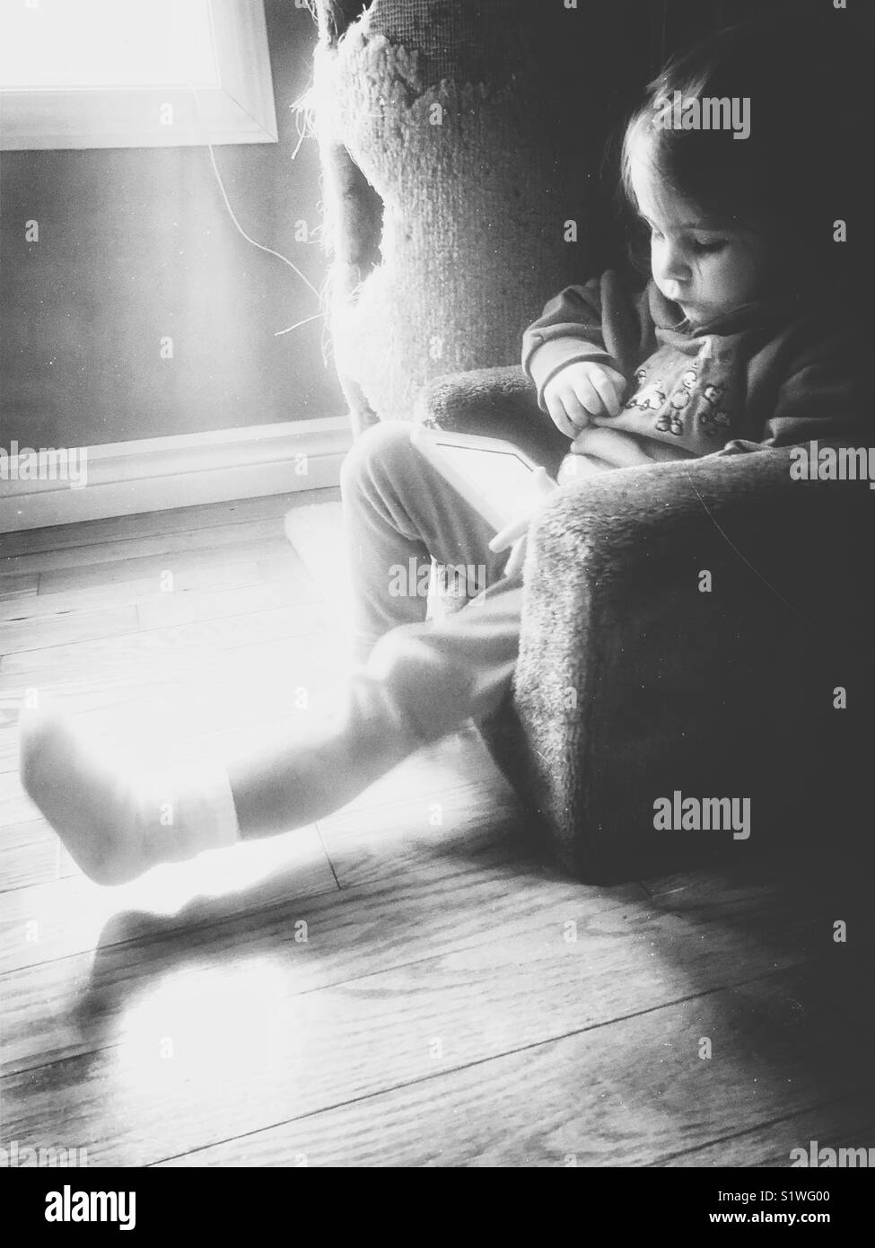 Black and white image of toddler girl sitting in child’s plush chair beside a window while playing on a tablet - Smartphone Captured Stock Image