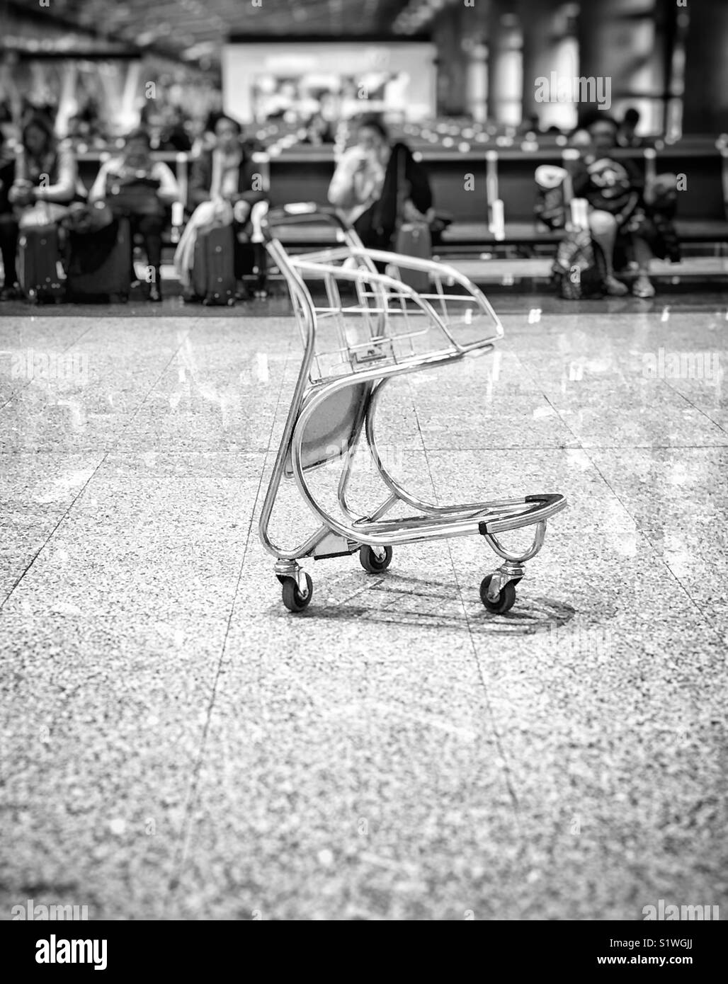 Empty baggage trolley in departure lounge of airport with rows of seated passengers in the background. Photo in monochrome Stock Photo