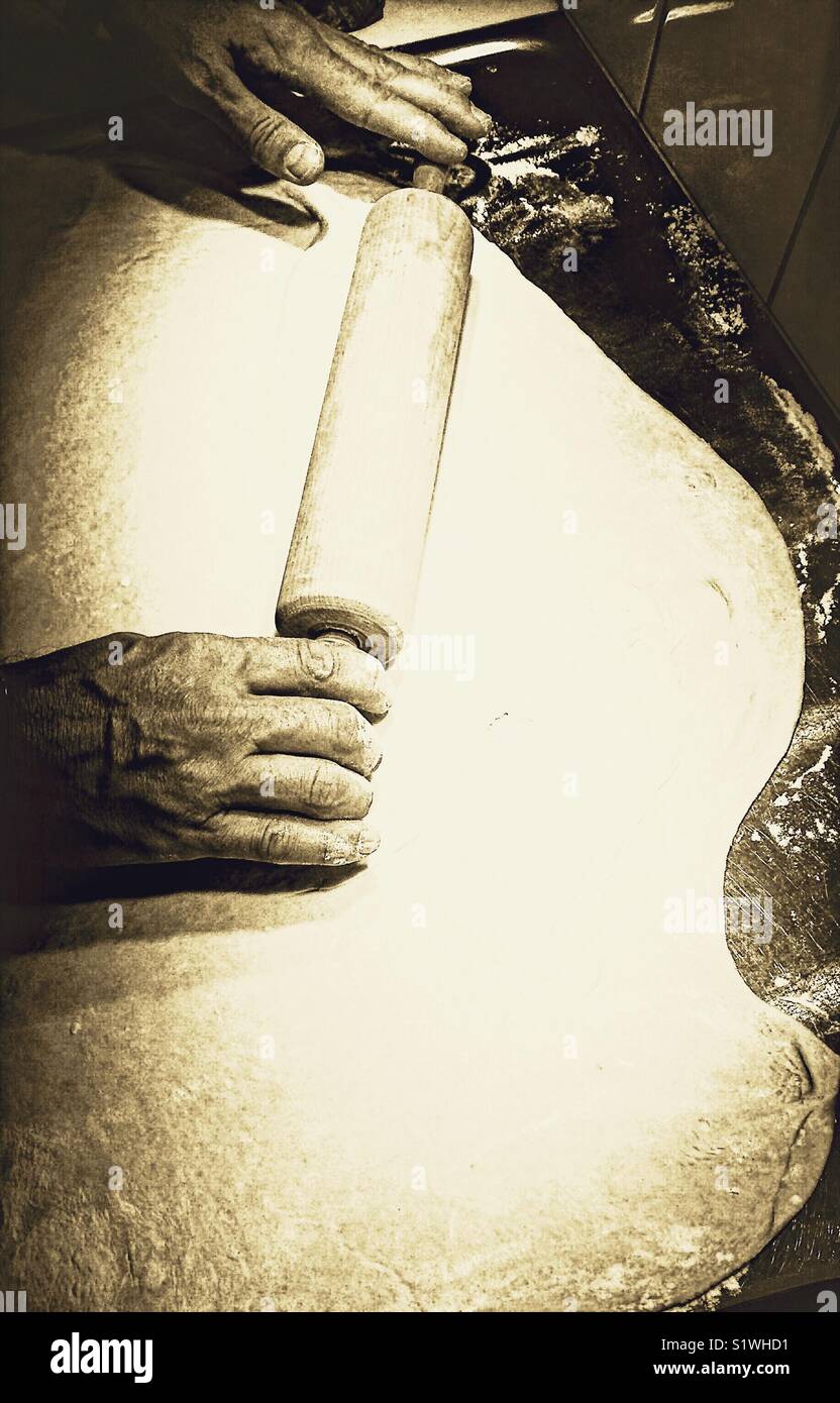 Male baker rolling out dough for pizza with wooden rolling pin on metal surface - Smartphone Captured Stock Image
