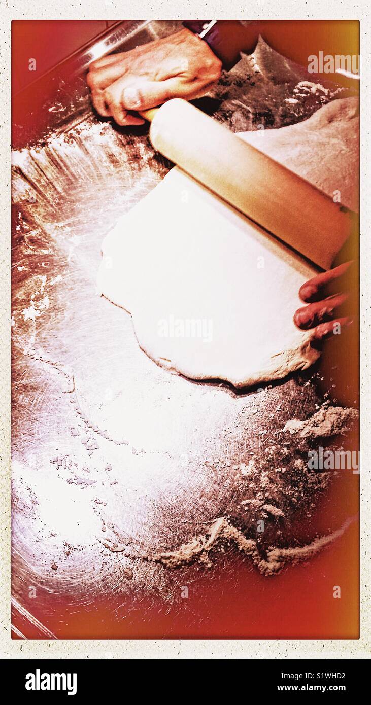 Male baker rolling out dough for pizza with wooden rolling pin on metal surface - Smartphone Captured Stock Image