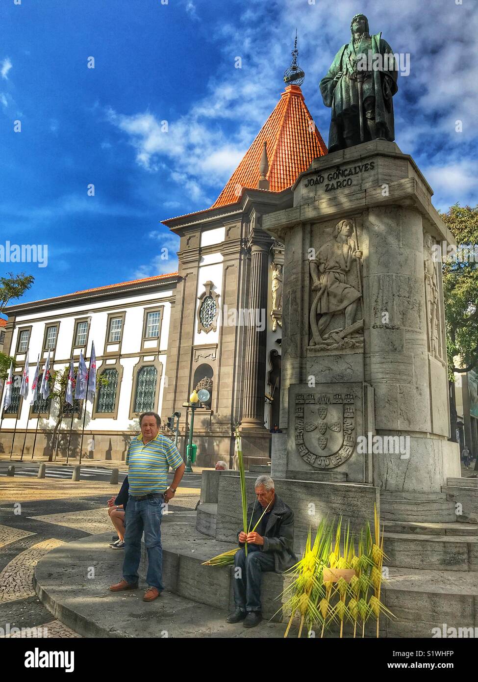 Traditional craftsman weaving, sitting by the statue of Zarco  with the iconic Bank of Portugal in the background. Funchal, Madeira, Portugal - Smartphone Captured Stock Image