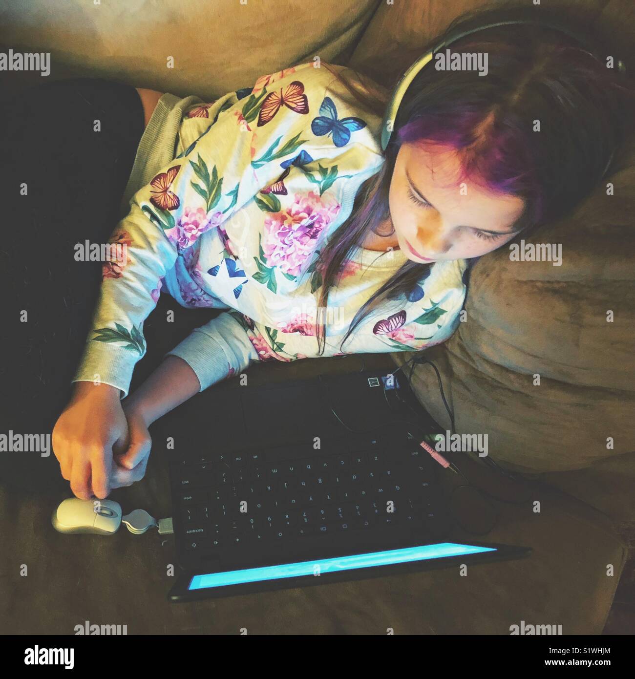 Girl with lavender hair using Chromebook with headphones on a cozy brown chair - Smartphone Captured Stock Image