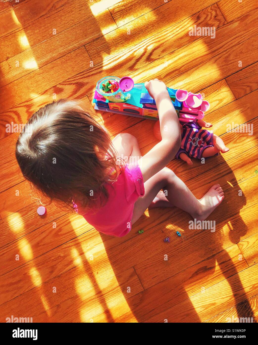 Toddler girl playing with toys on wooden floor with sunlight making a blinds pattern over her Stock Photo