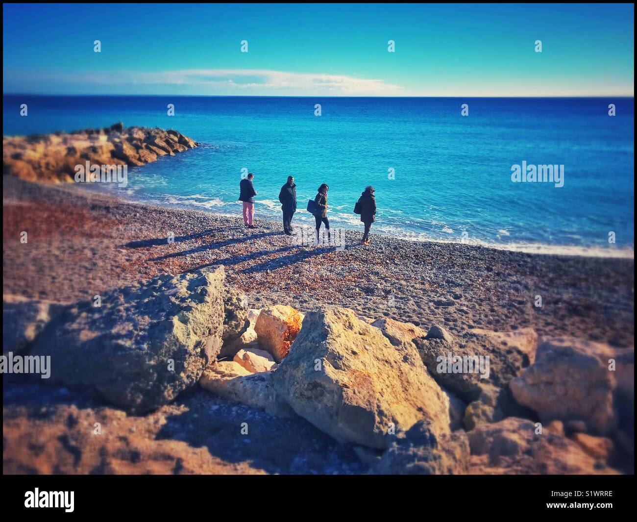4 people on a beach next to the Mediterranean Sea. Despite the blue sky & sunshine, it is winter hence the warm jackets. Photo Credit © COLIN HOSKINS. - Smartphone Captured Stock Image