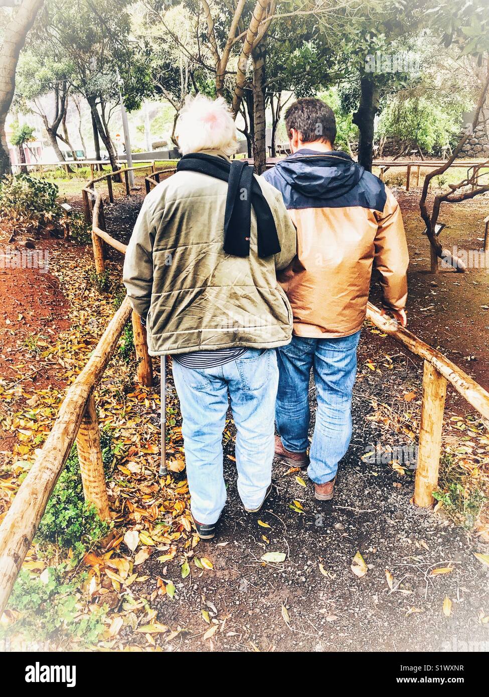 Senior man walking with a stick and with the aid of a younger man, helping him. Back view. - Smartphone Captured Stock Image
