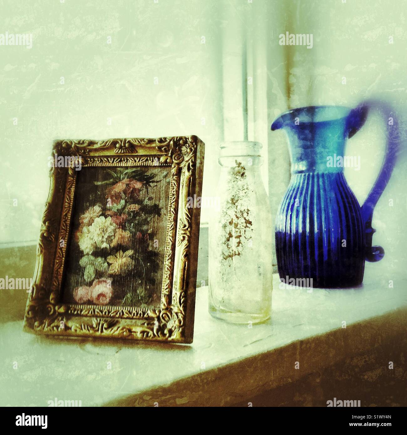 Nostalgic Shabby chic still life on a windowsill with antique blue glass pitcher, dusty glass milk bottle and floral painting in gold frame Stock Photo