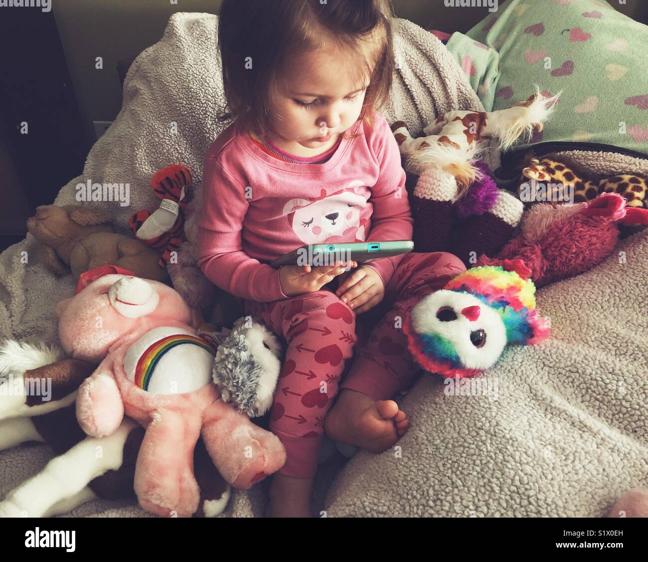 Toddler girl in pink heart pyjamas playing on tablet surrounded with stuffed toys - Smartphone Captured Stock Image