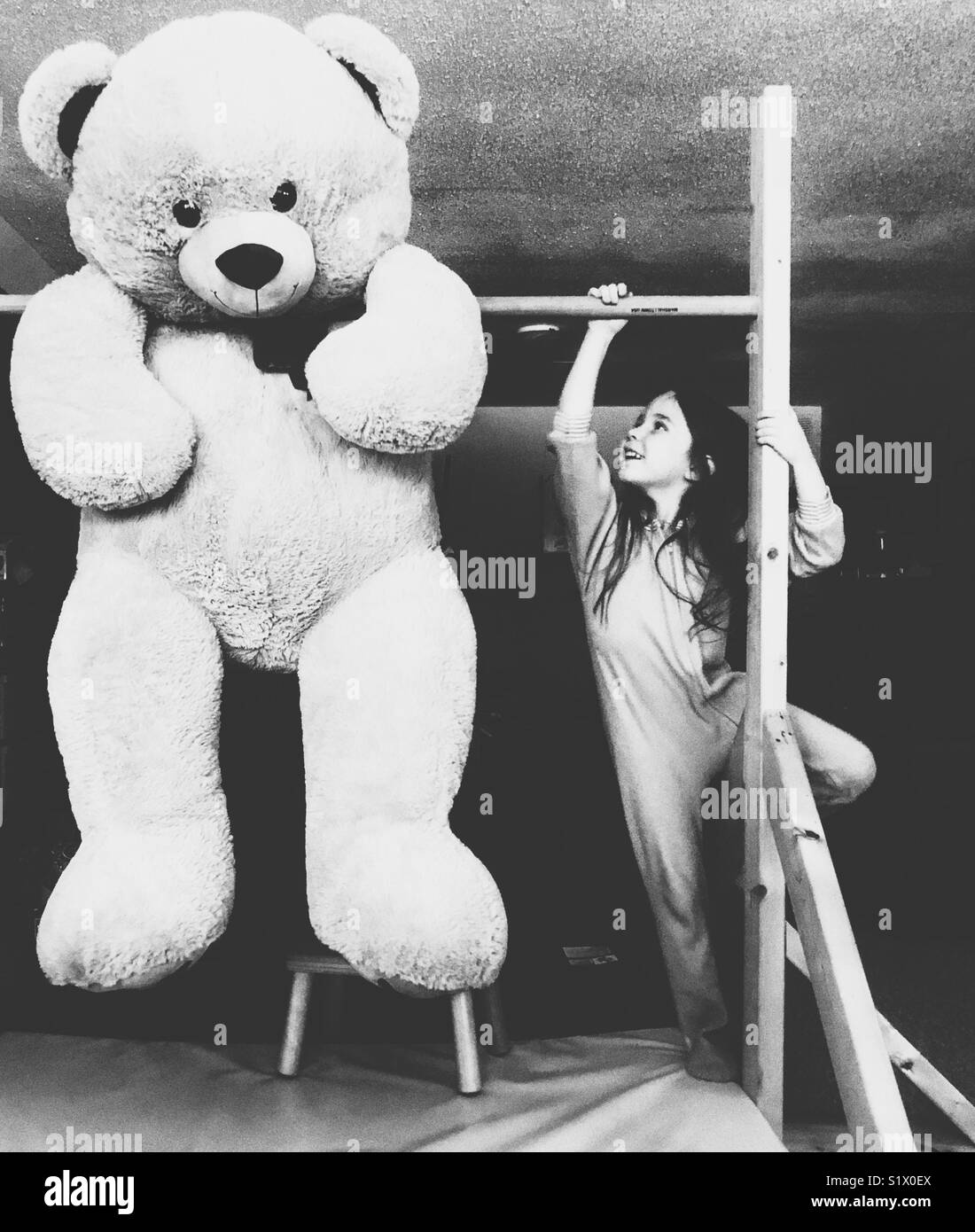Young girl with long hair in pyjamas climbing to reach giant oversized teddy bear hanging on wooden gymnastics bar Stock Photo