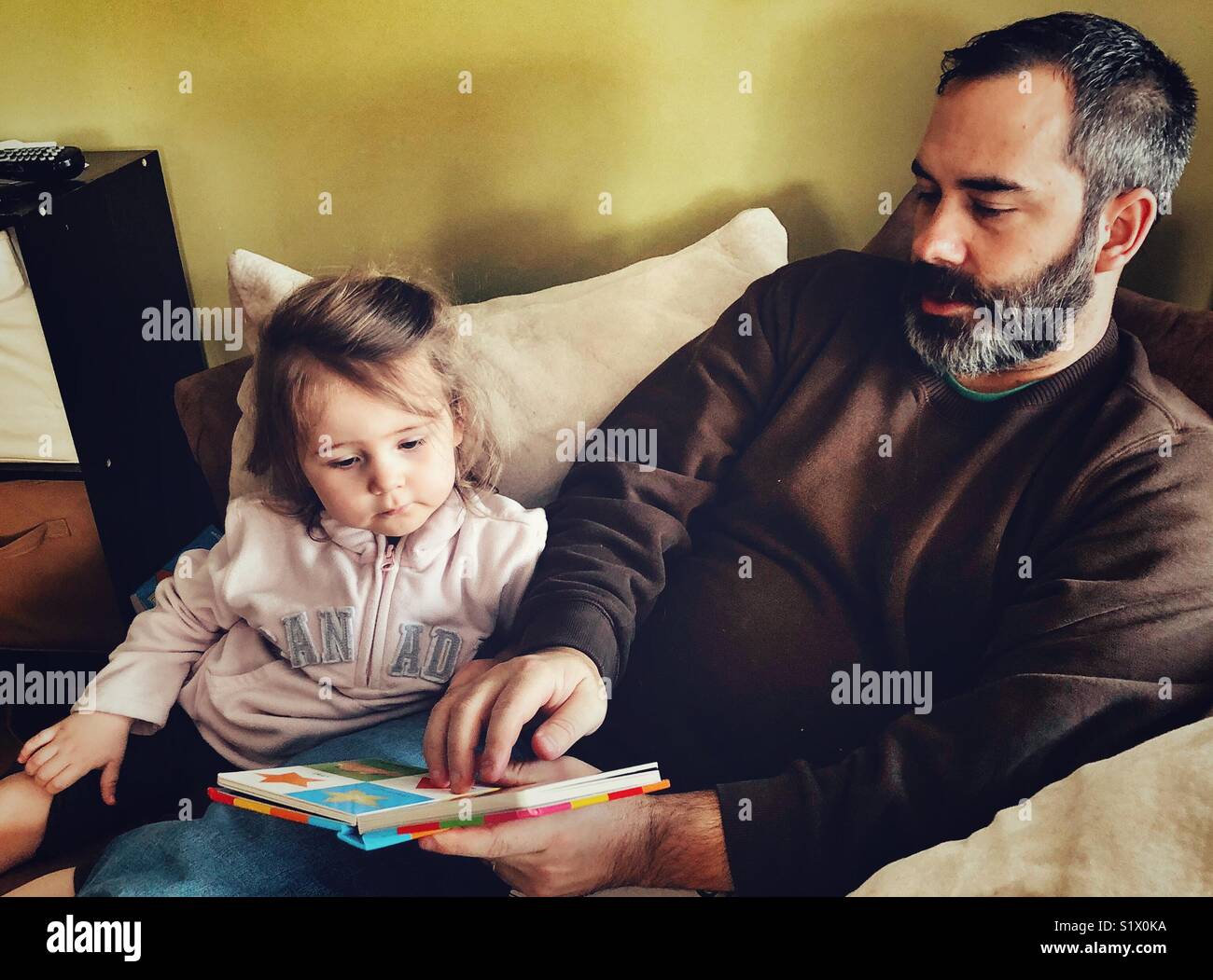 Middle aged father with beard reading a board book to toddler daughter - Smartphone Captured Stock Image