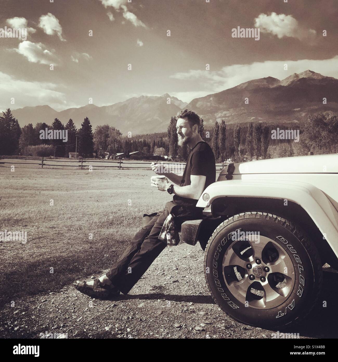A man sits on the front of a 4x4 eating food with a great view of the mountains behind - Smartphone Captured Stock Image