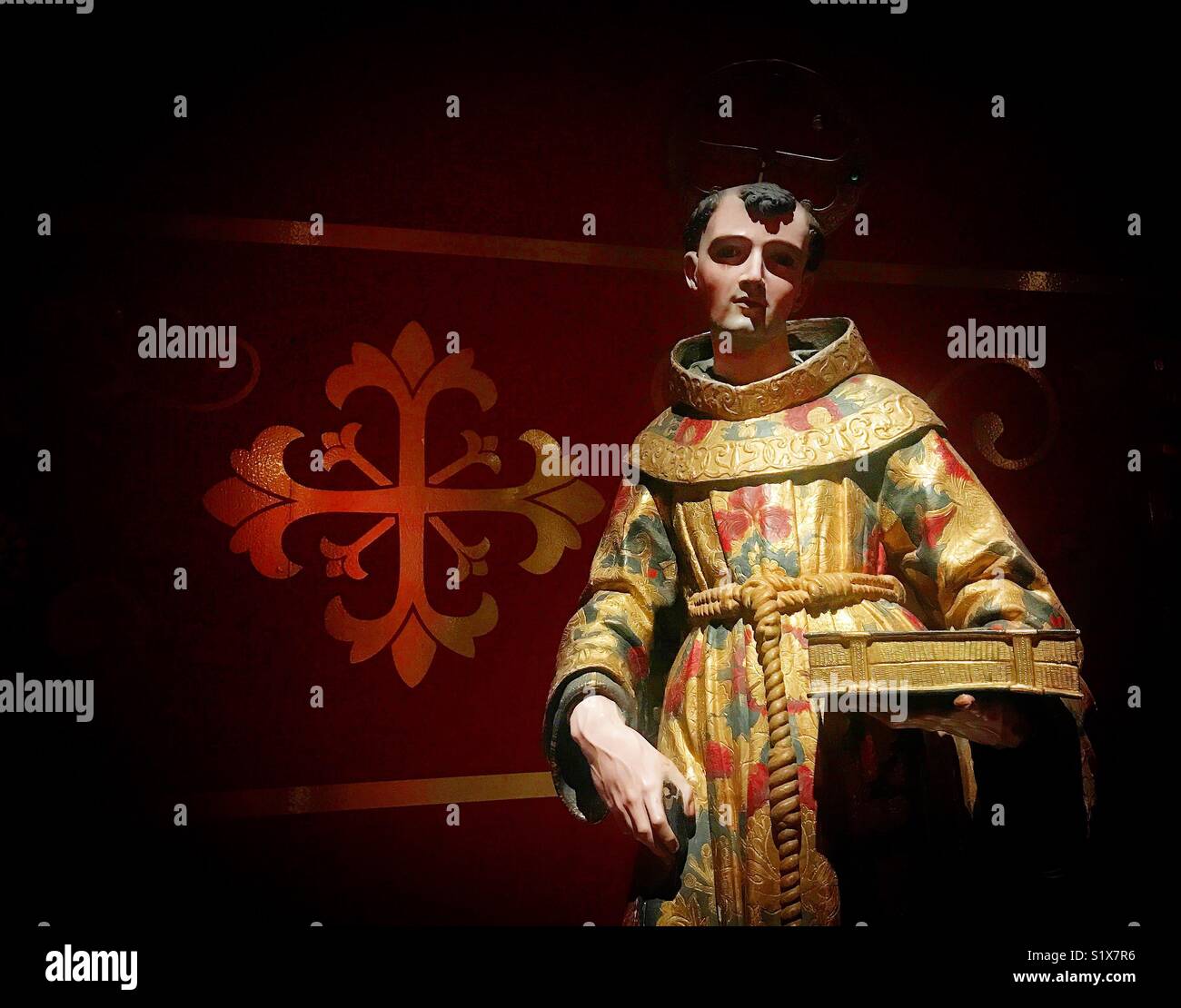 A sculpture of Saint Anthony of Padua in front of a red cross is displayed in Galeria Tesoros de la Catedral museum in Puebla de los Angeles, Mexico - Smartphone Captured Stock Image