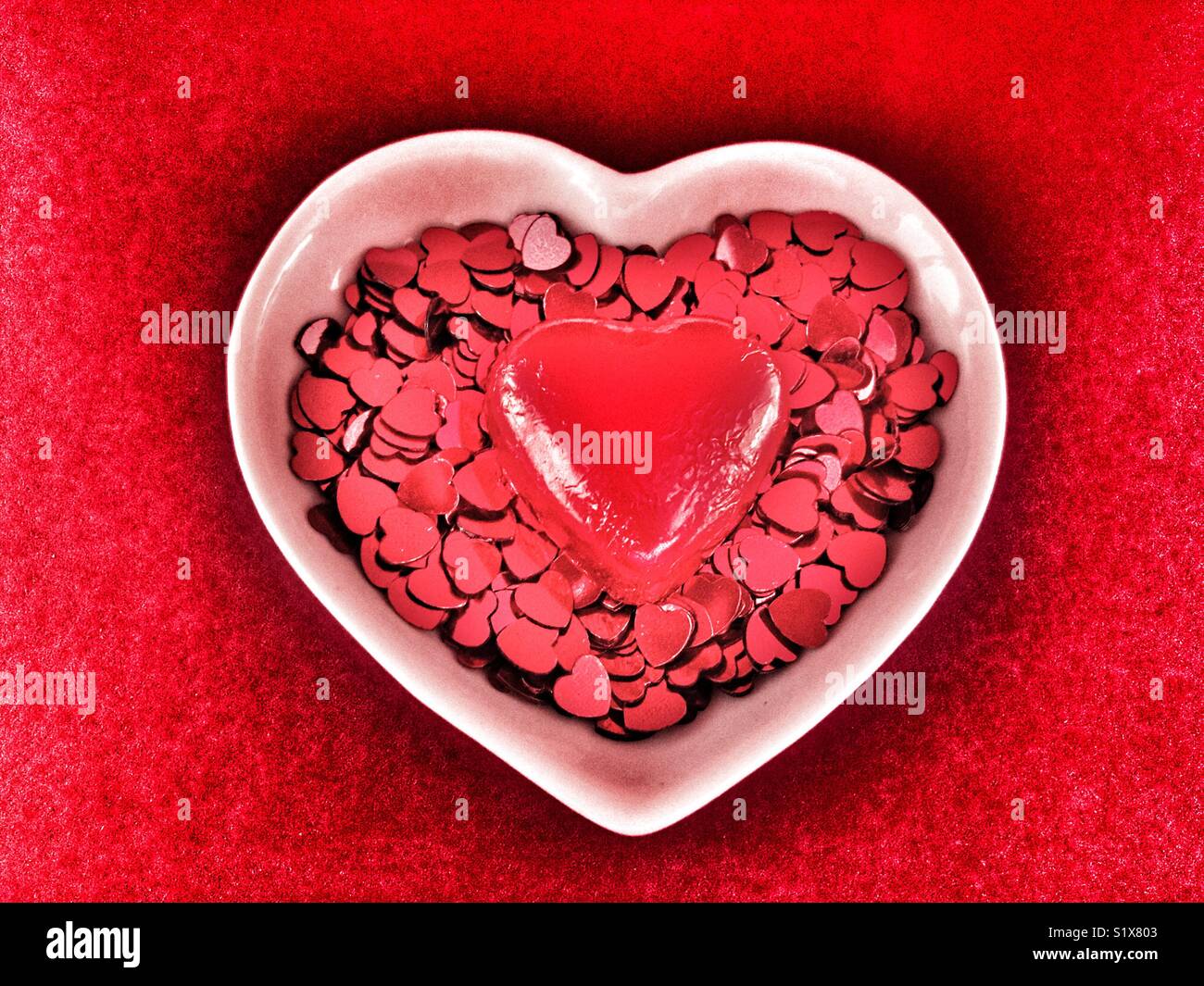 Valentine’s Day, Red heart sprinkles and a red chocolate heart  in a white heart shaped ceramic dish, red background - Smartphone Captured Stock Image