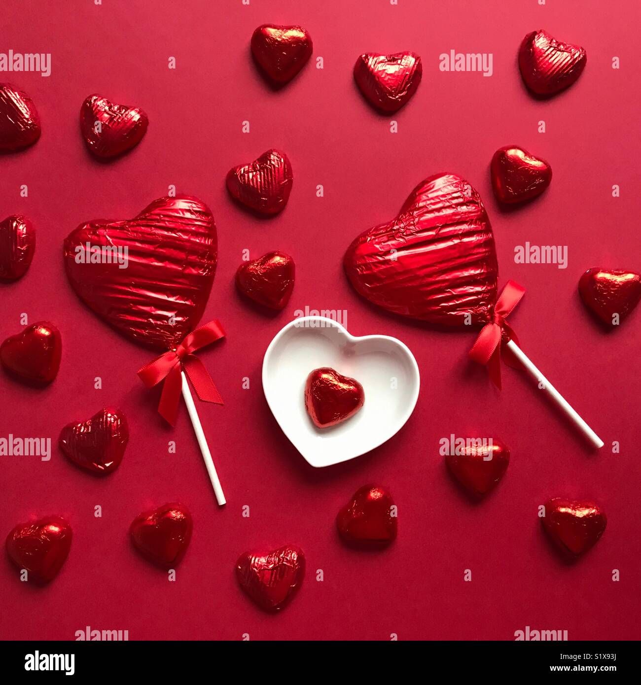 Flatlay photography. Heart shaped chocolate lollipops and heart shaped chocolate hearts wrapped in red foil, on red background - Smartphone Captured Stock Image