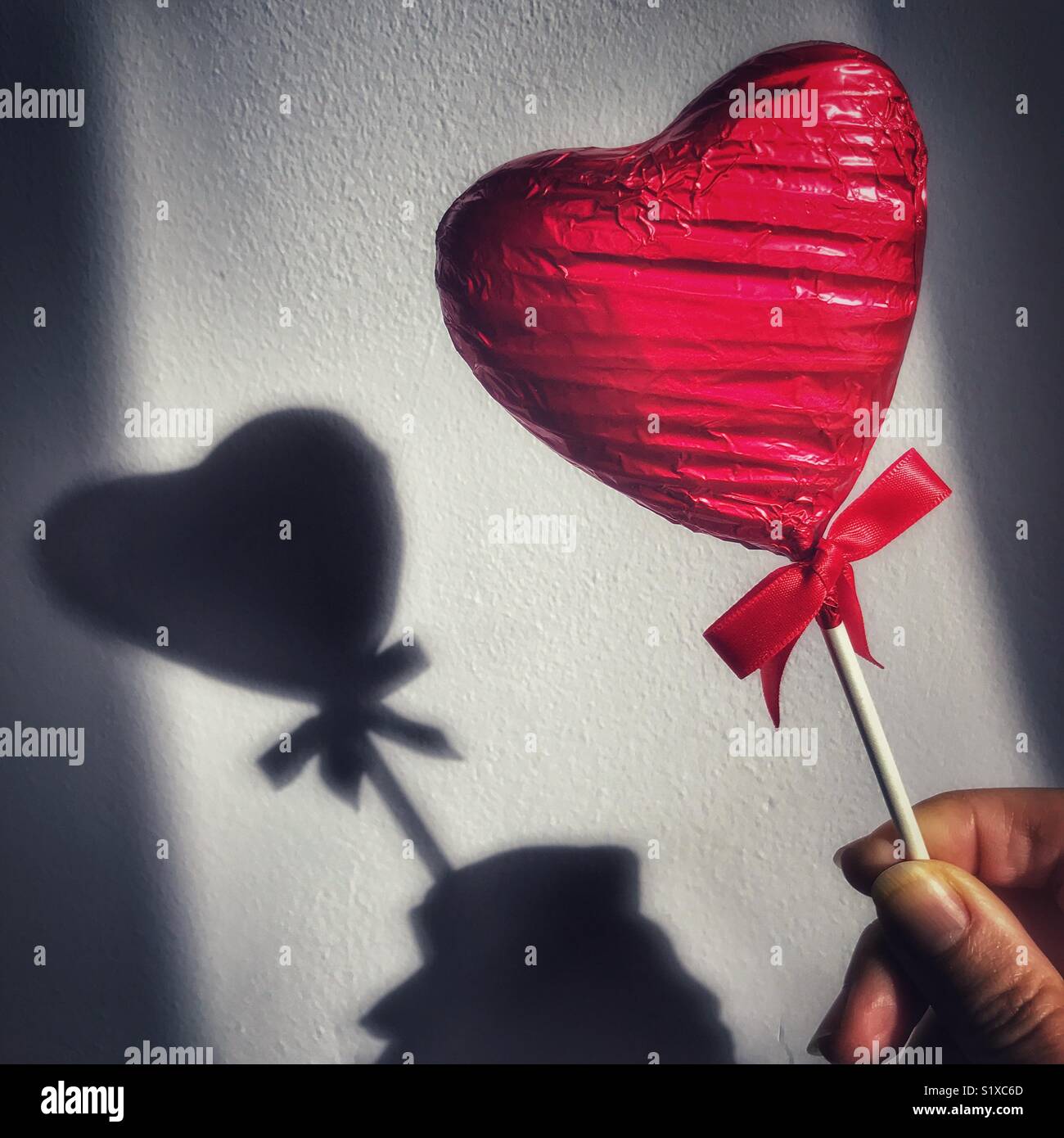 Heart shaped, foil wrapped, chocolate lollipop, in a woman’s hand with shadow on the wall - Smartphone Captured Stock Image