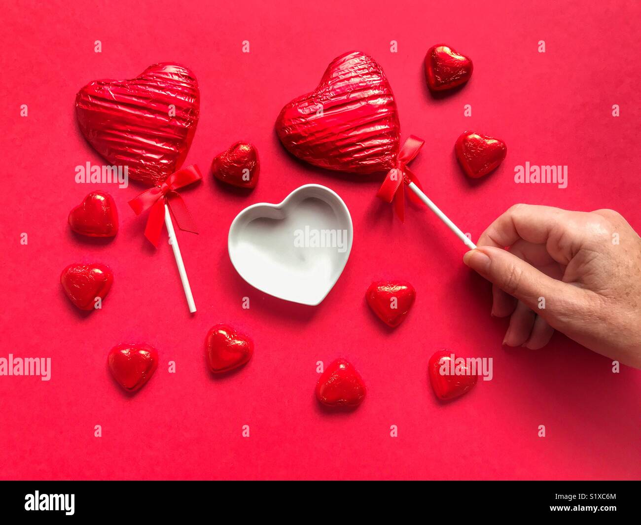 Heart shaped, foil wrapped, chocolate lollipop, in a woman’s hand with chocolate hearts, lollipop and ceramic heart shape dish - Smartphone Captured Stock Image