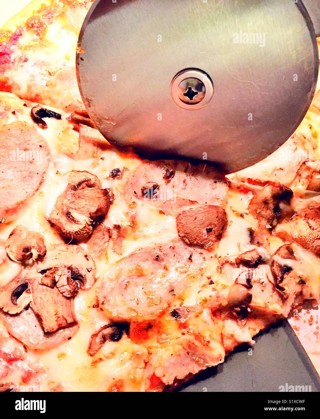 Metal pizza cutting wheel slicing through fresh hot sausage and mushroom pizza - Smartphone Captured Stock Image