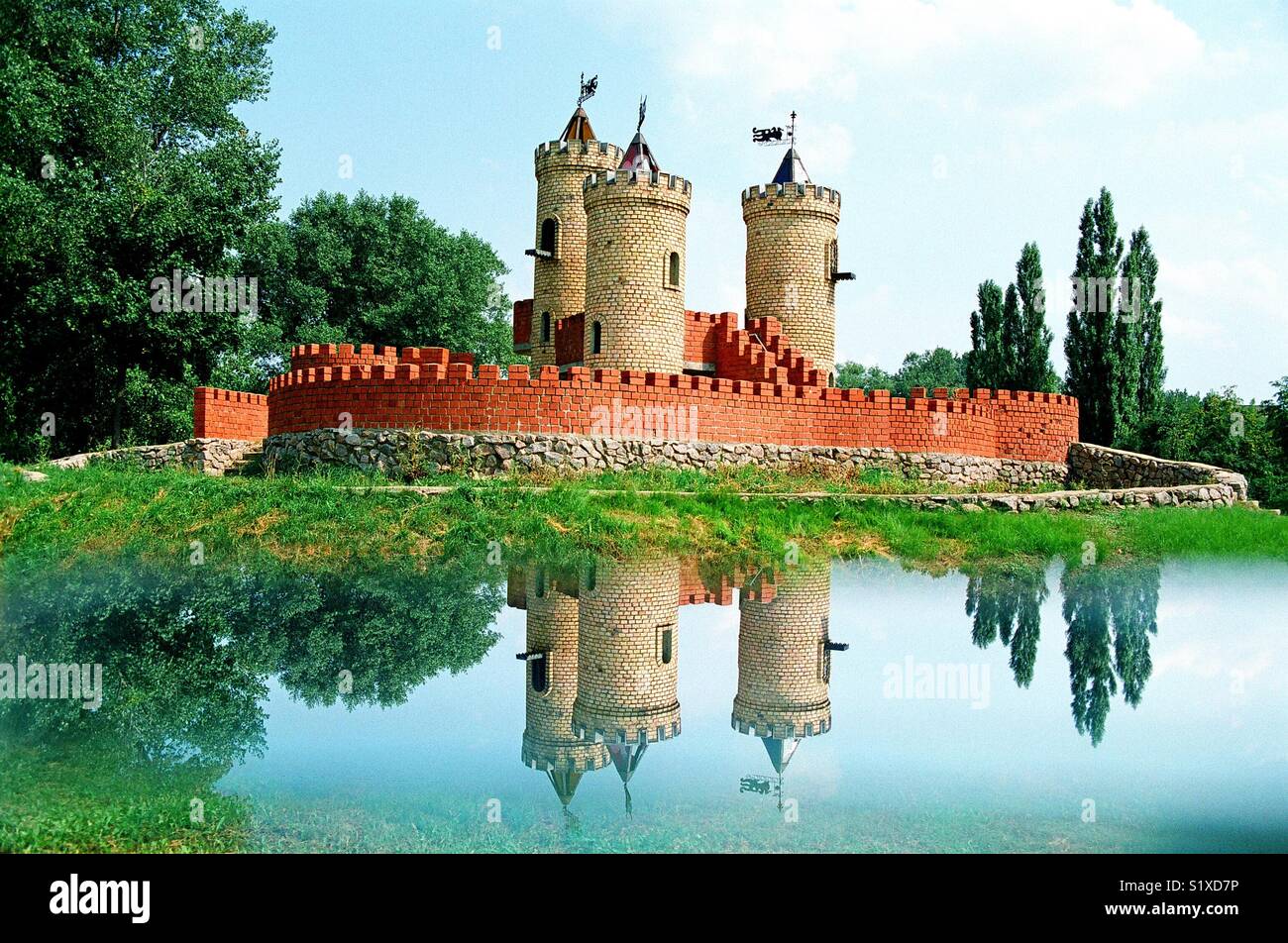 Fairy-tale like dreamy castle medieval red brick kids playground fortress reflected in water surface of the moat in children's park on a sunny summer day in Sumy, Ukraine Stock Photo