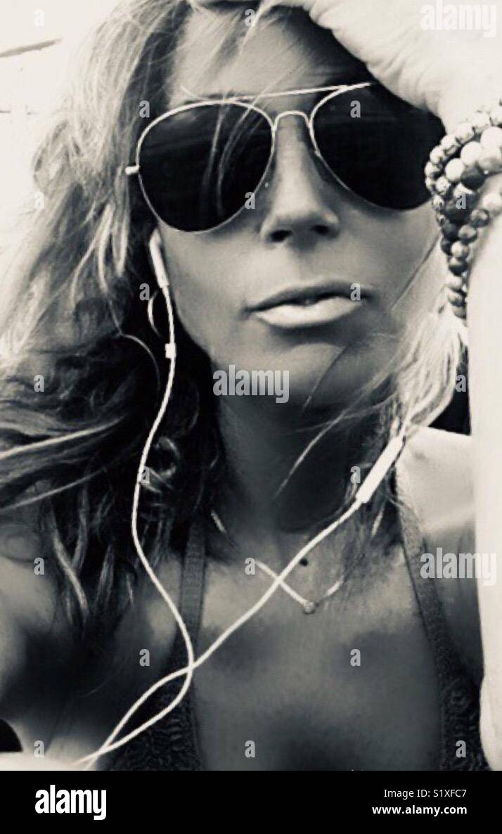 Black and white portrait of a woman outside soaking up the sun with sunglasses and earphone listening to music Stock Photo