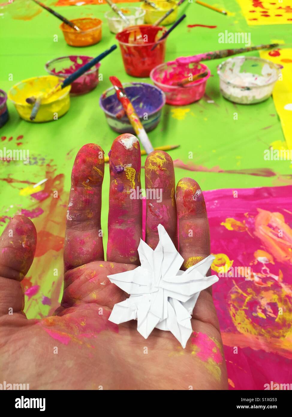 Origami flower made by a child on art teacher's hand covered with paint in front of table with colorful paints - Smartphone Captured Stock Image