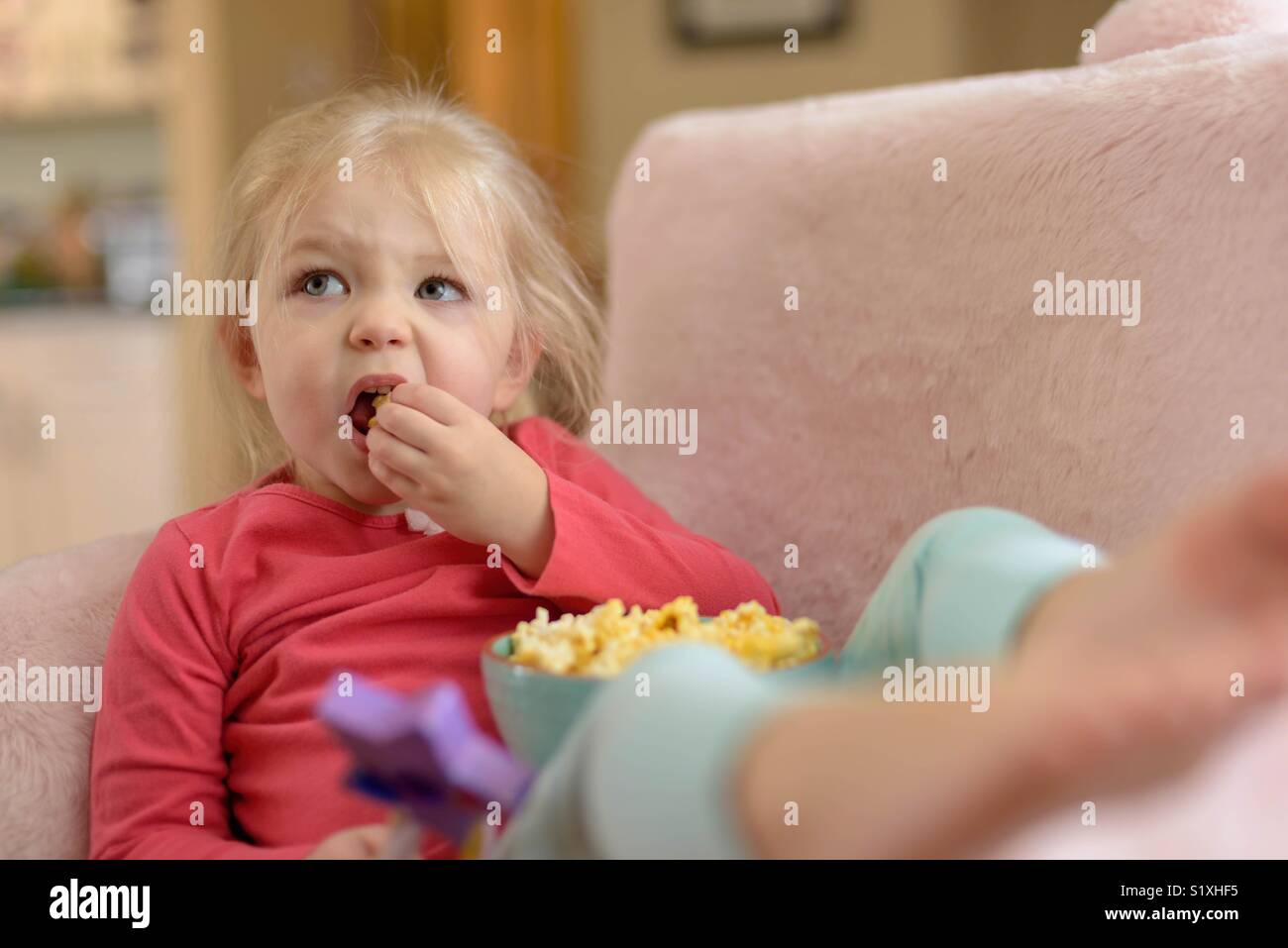Cute girl making funny face while eating popcorn and watching TV Stock Photo