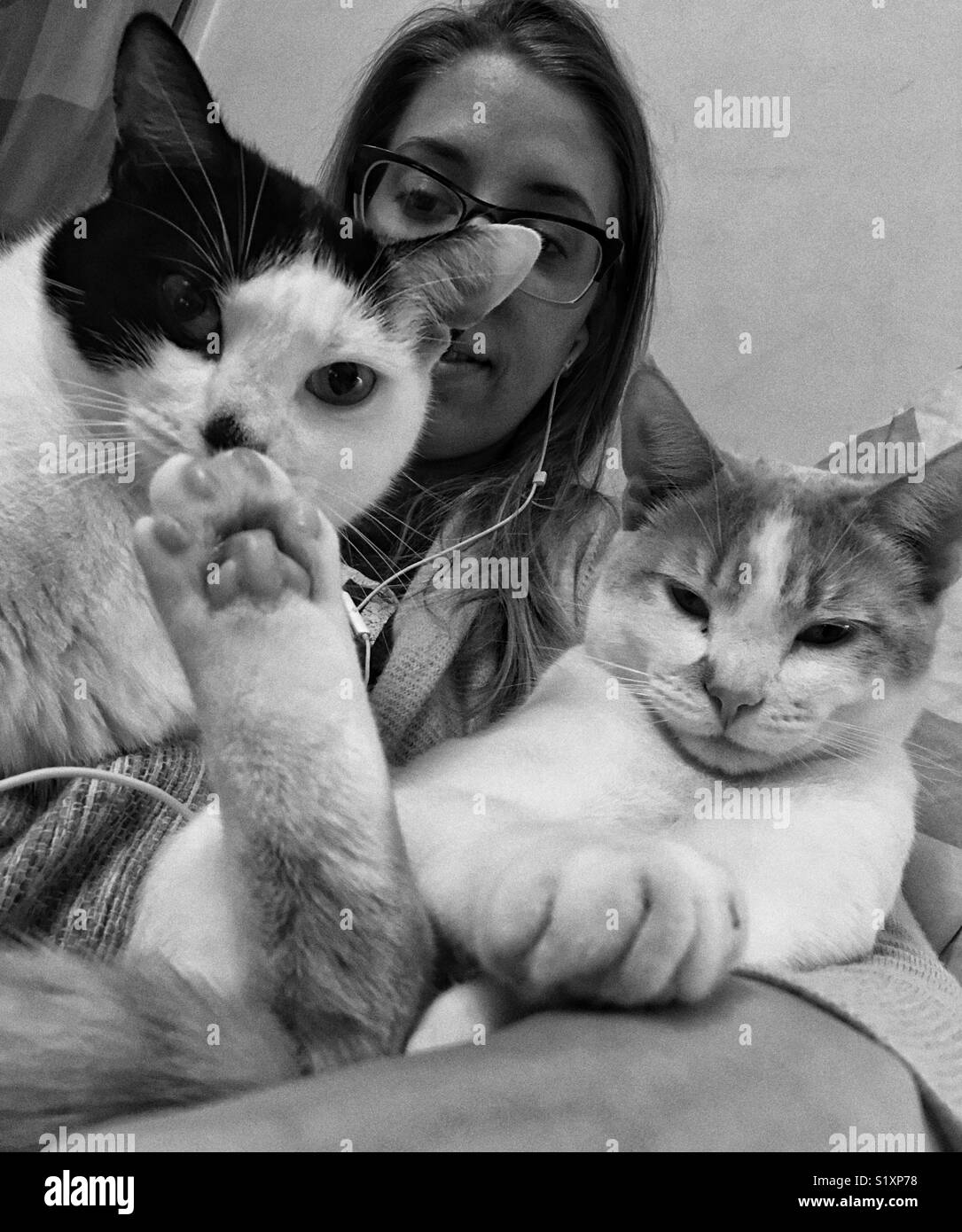 A black and white photograph of a woman sitting holding two cats while she listens to music on a Saturday evening Stock Photo