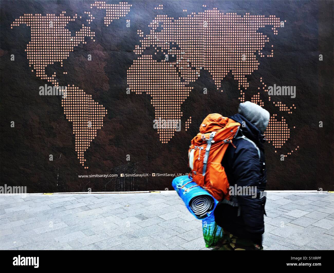 A man wearing a back pack walks past a map of the world in London. Stock Photo
