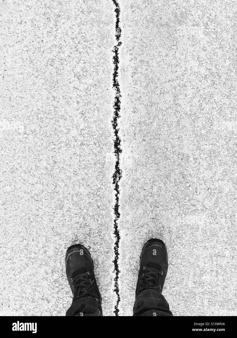 First-person perspective of a man's feet standing either side of a large vertical crack in the road. Stock Photo