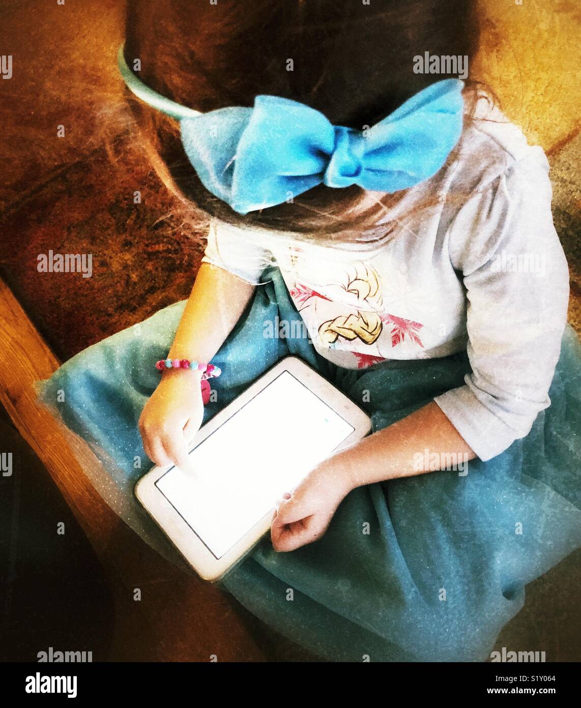 Young girl wearing blue dress and hair bow sitting cross legged on floor with a tablet Stock Photo