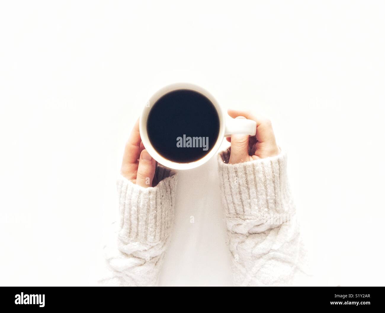 Looking down from above onto a white background with a girl’s hands holding a cup of black coffee and wear a warm arran sweater - Smartphone Captured Stock Image