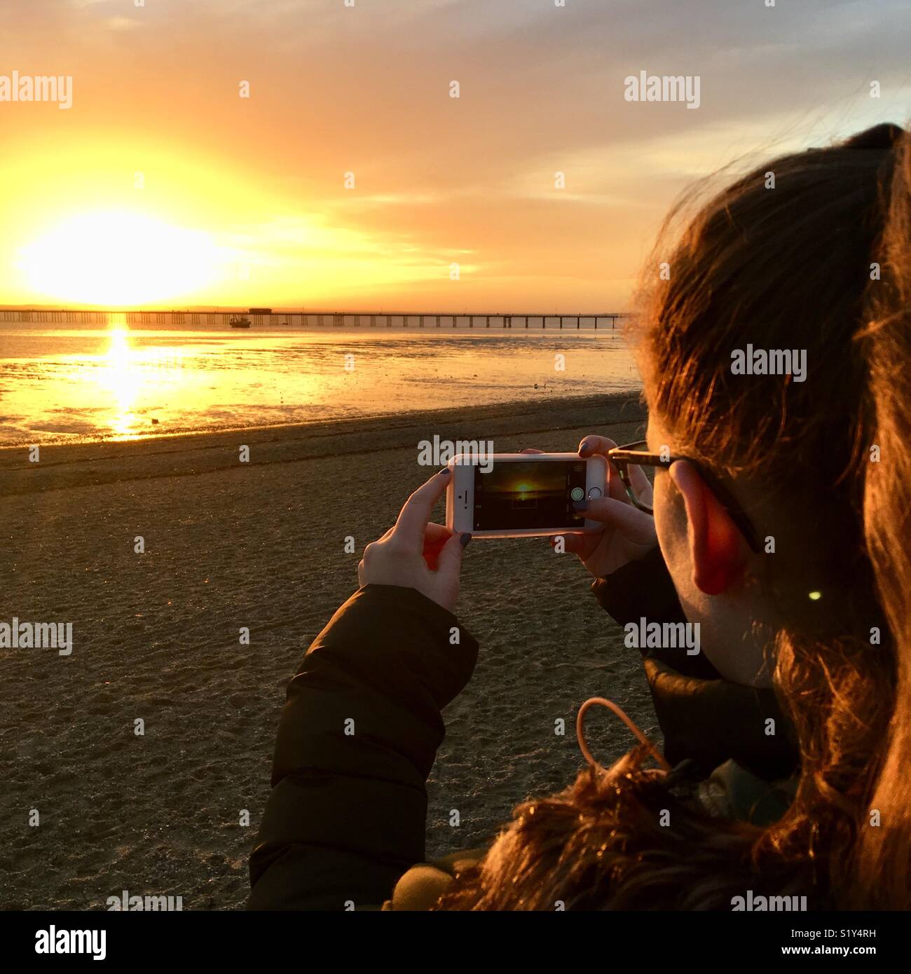 Young girl takes iPhone photo of a glowing sunset over Southend Pier, Essex, UK Stock Photo