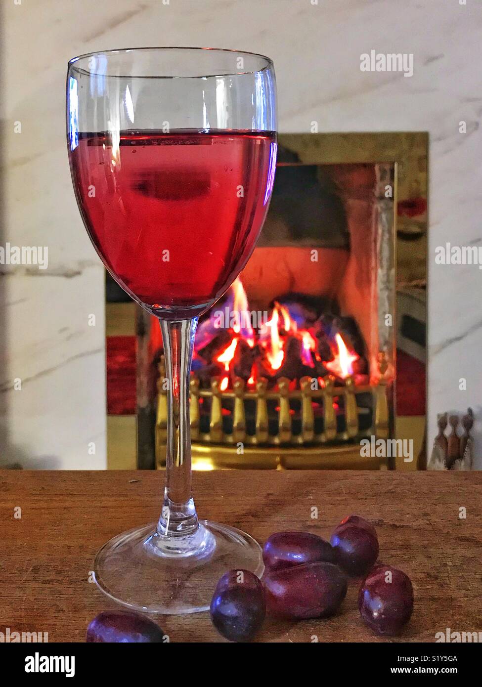 Still life of a glass of red wine and red grapes on a wooden table in front of a coal fire with brass surrounds - Smartphone Captured Stock Image