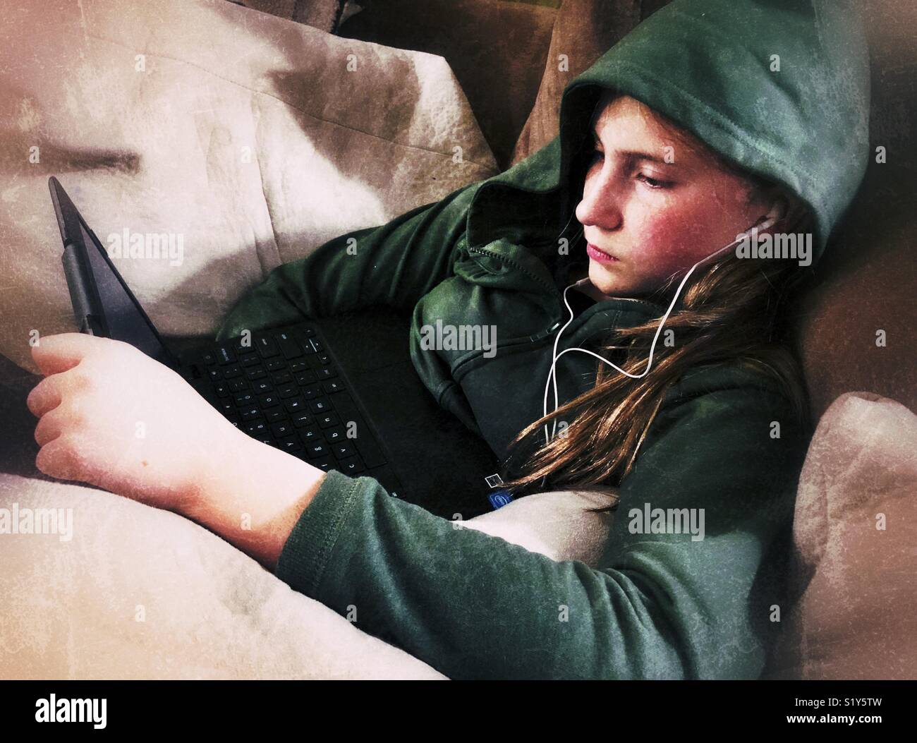 Teen girl wearing green hoodie with hood on using Chromebook laptop with white earbuds - Smartphone Captured Stock Image