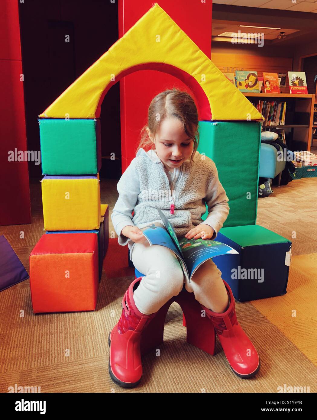 Girl sitting on stool in children’s area of public library reading a book - Smartphone Captured Stock Image