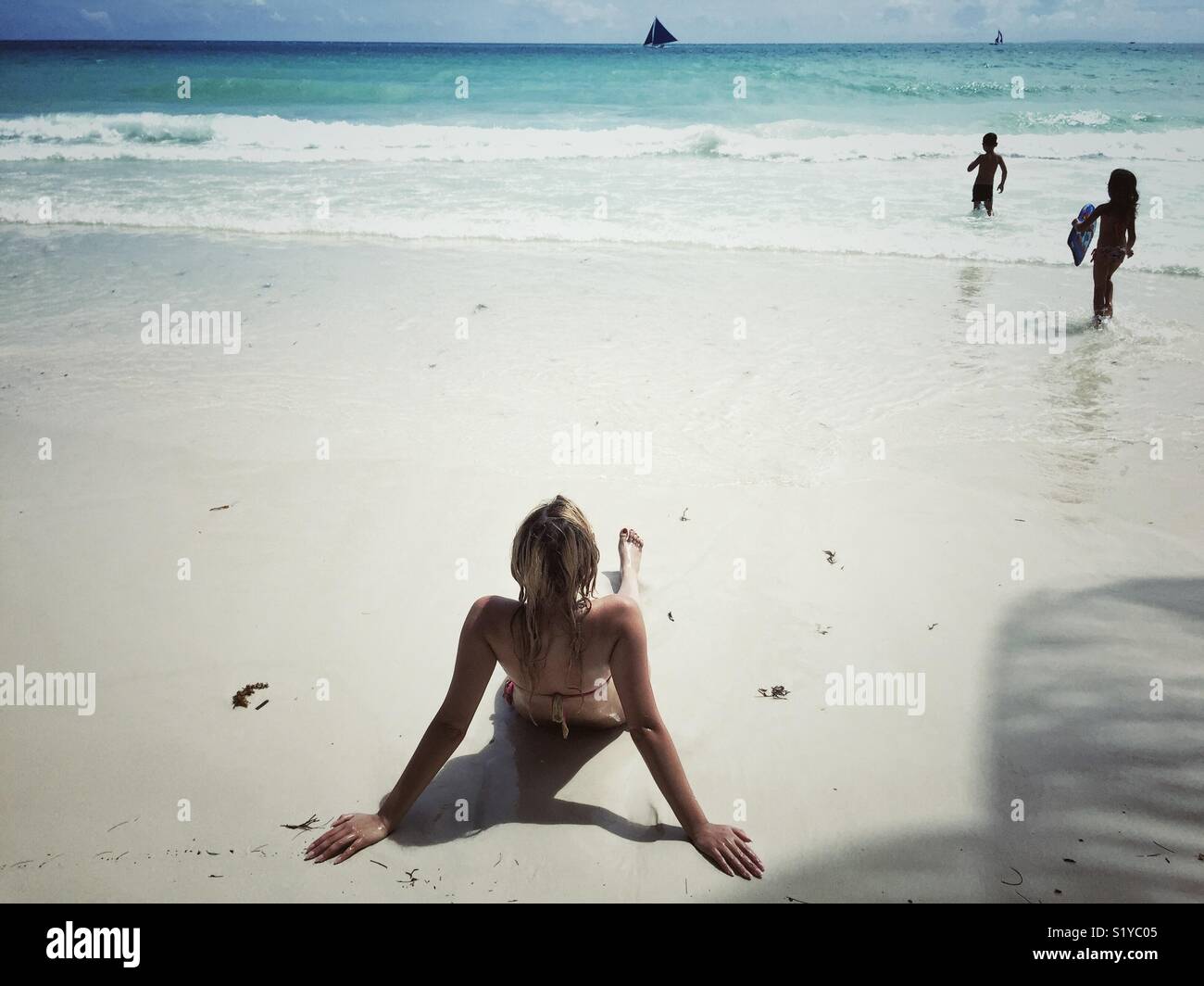 Young woman on white sand beach in bikini watching kids playing in water Stock Photo