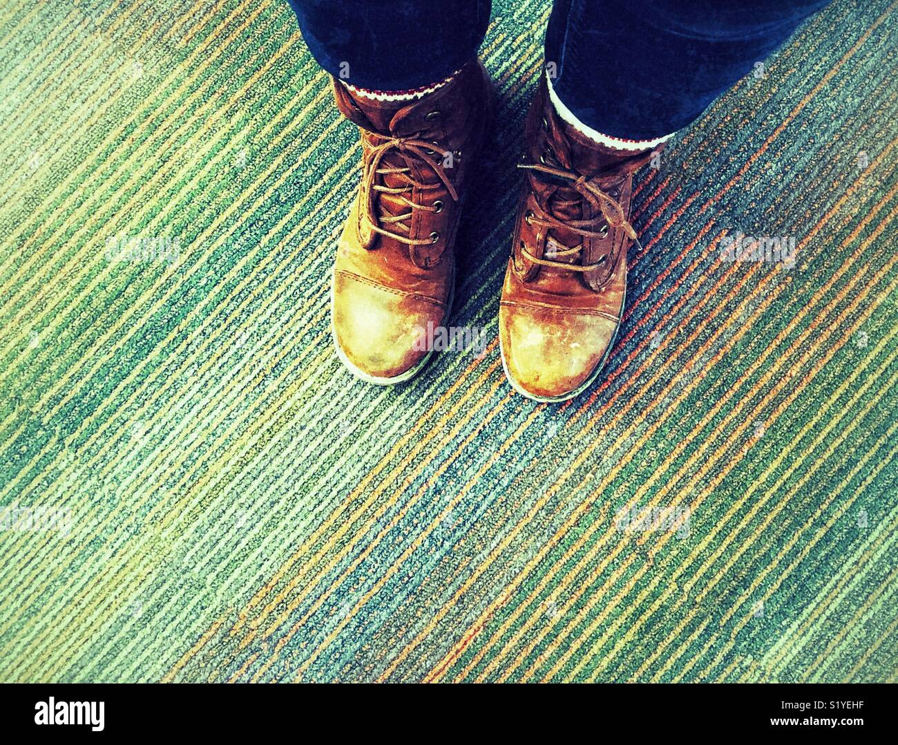 Looking down at person’s feet wearing scuffed, old, brown work boots on a multicoloured carpet - Smartphone Captured Stock Image