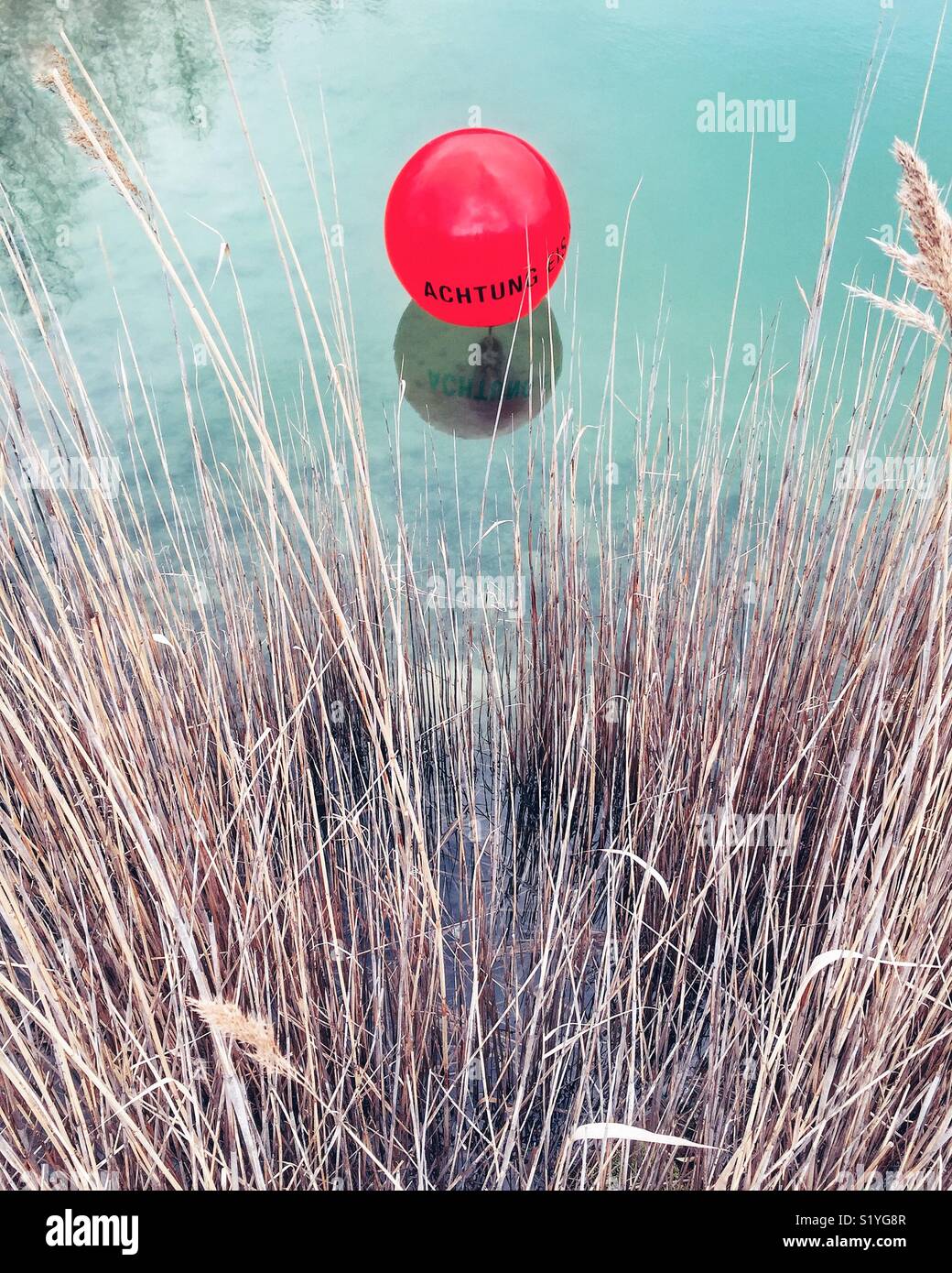 A red balloon with the message 'Achtung' floating on the surface of a pond, with reeds and weeds Stock Photo
