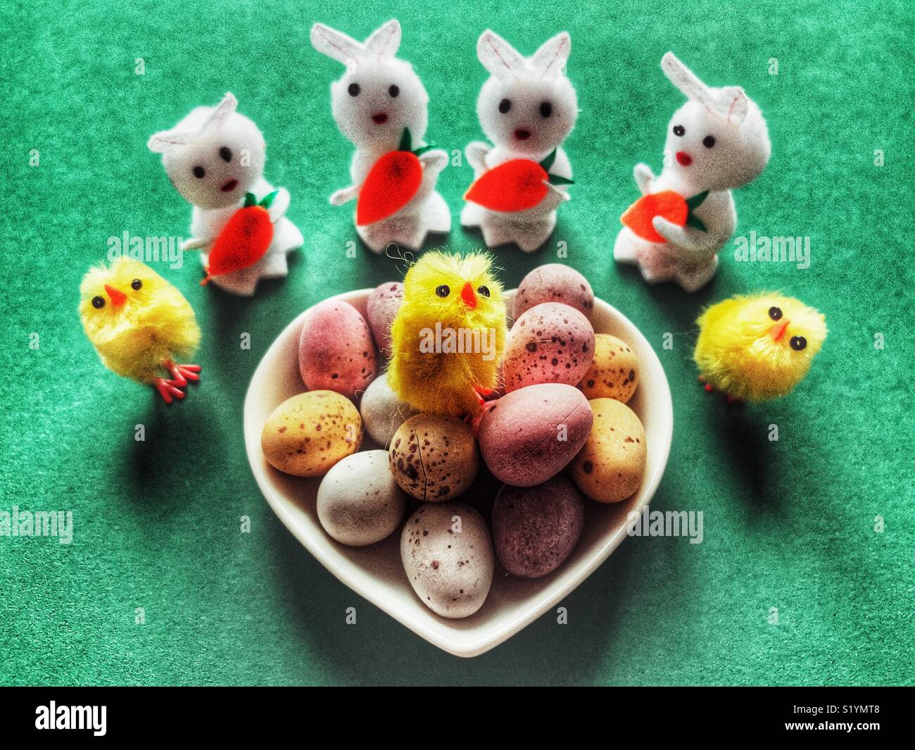 Sugar coated mini chocolate Easter eggs in a heart shaped dish, with mini toy Easter bunnies  and Easter chicks on a green background - Smartphone Captured Stock Image