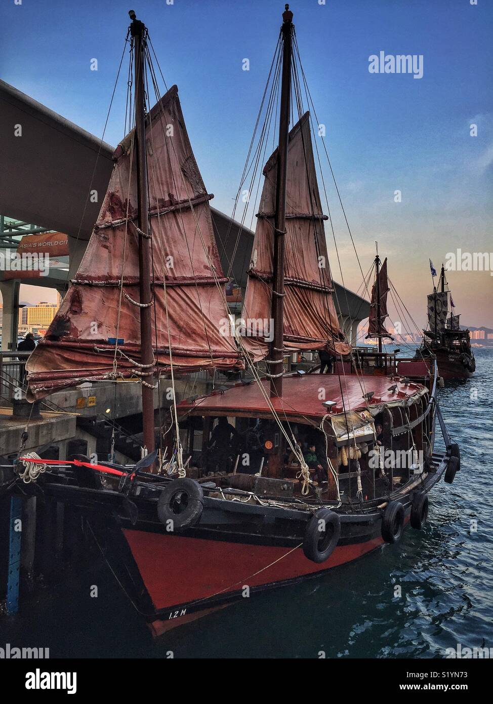 The ‘Duk Ling’, a replica Chinese junk, used for sightseeing cruises on Victoria Harbour, picking up tourists at Pier 9, Central, Hong Kong Island at sunset - Smartphone Captured Stock Image