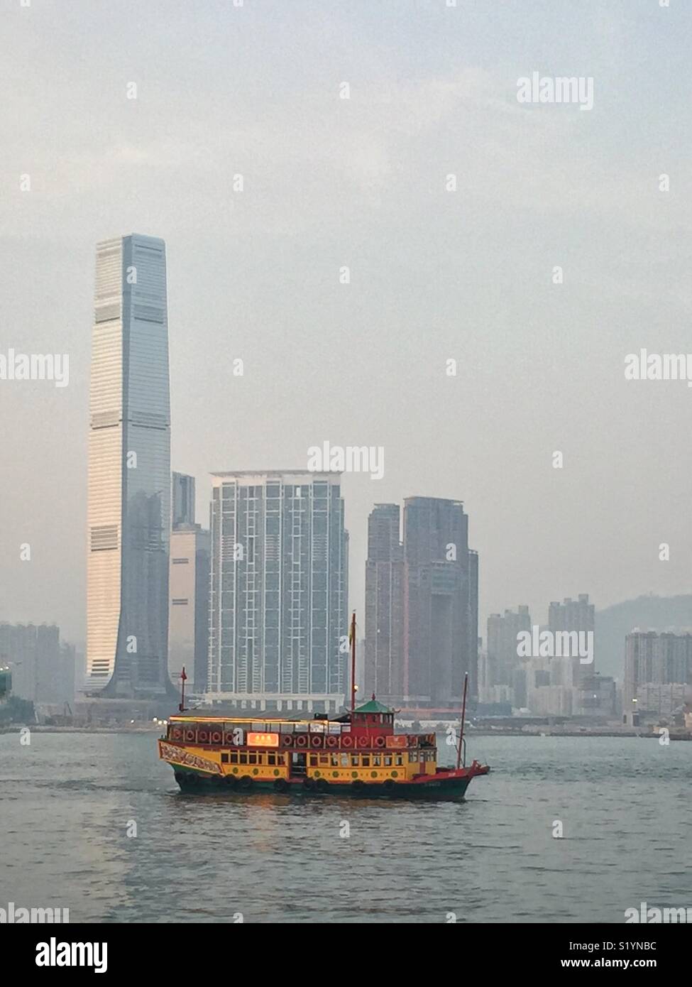 The ‘Wing On Travel’, a double decker Chinese junk, carrying tourists on a sightseeing cruise on Victoria Harbour at twilight, in front of the ICC, Hong Kong’s tallest skyscraper, in West Kowloon - Smartphone Captured Stock Image