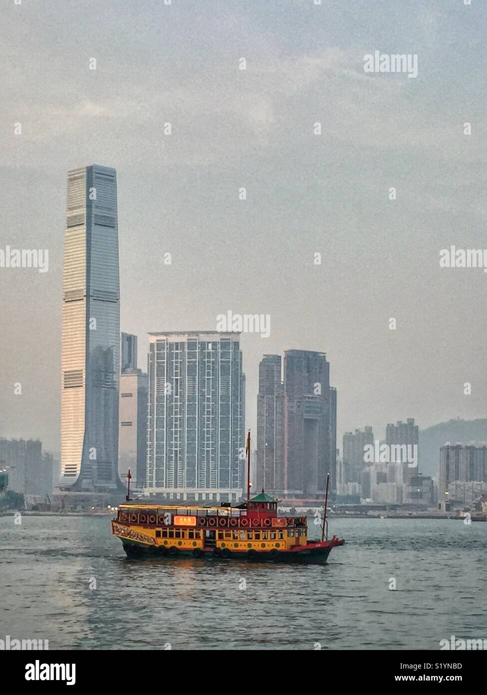 The ‘Wing On Travel’, a double decker Chinese junk, carrying tourists on a sightseeing cruise on Victoria Harbour at twilight, in front of the ICC, Hong Kong’s tallest skyscraper, in West Kowloon - Smartphone Captured Stock Image
