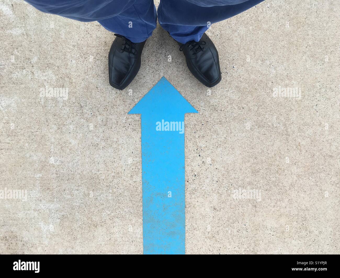 A painted blue arrow painted on the ground pointing to a man’s black shoes and blue pants. - Smartphone Captured Stock Image