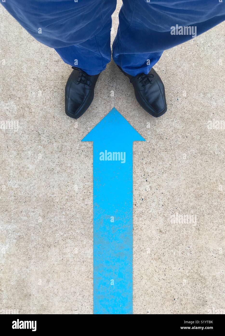 A blue arrow painted on the ground pointing to a man’s black shoes and blue pants. Stock Photo