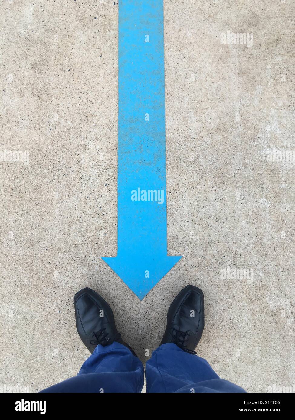 Blue arrow painted on the ground pointing to a man’s black shoes and blue pants. - Smartphone Captured Stock Image