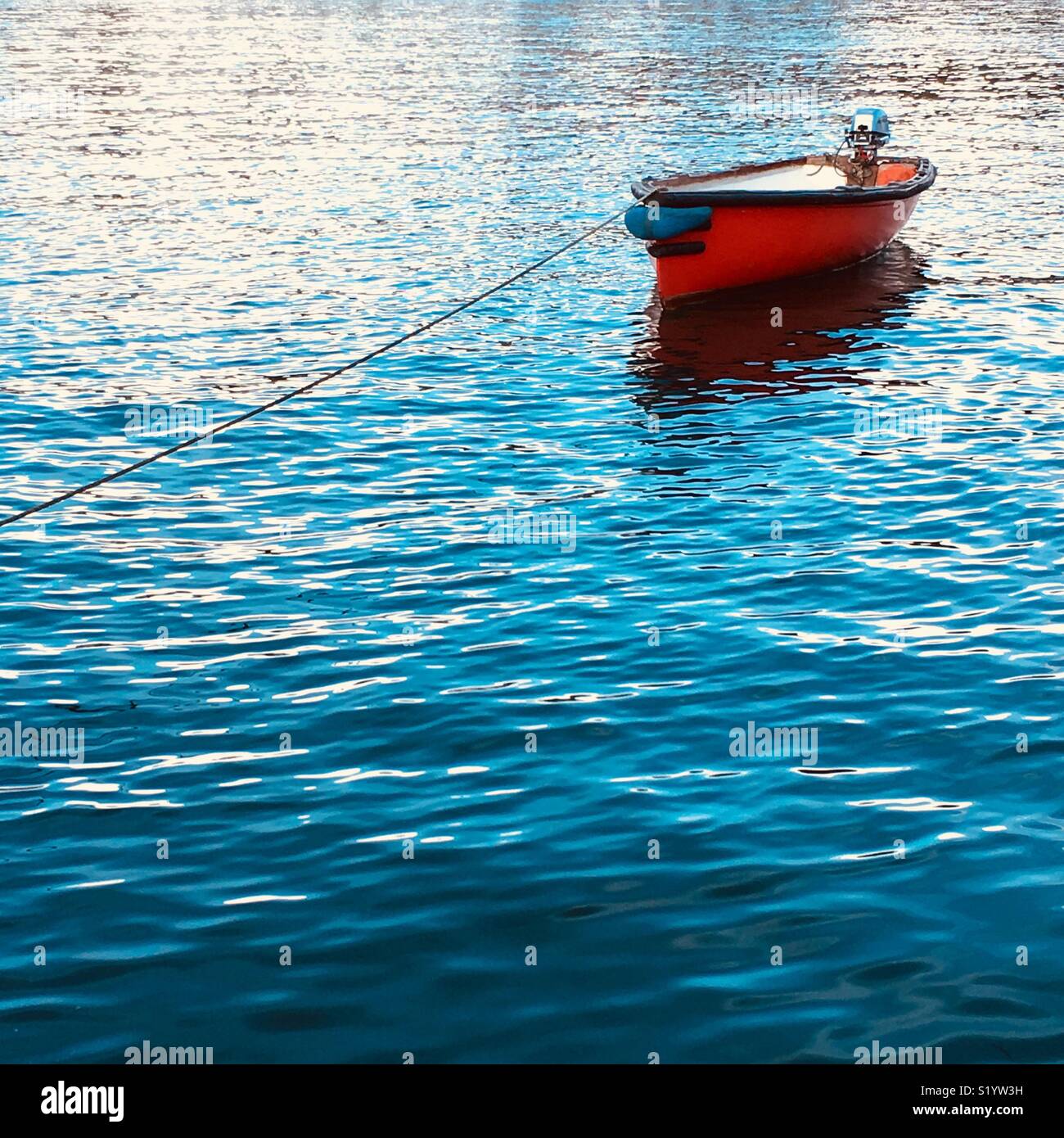 A red hulled open boat with an outboard motor tied up in the sea - Smartphone Captured Stock Image