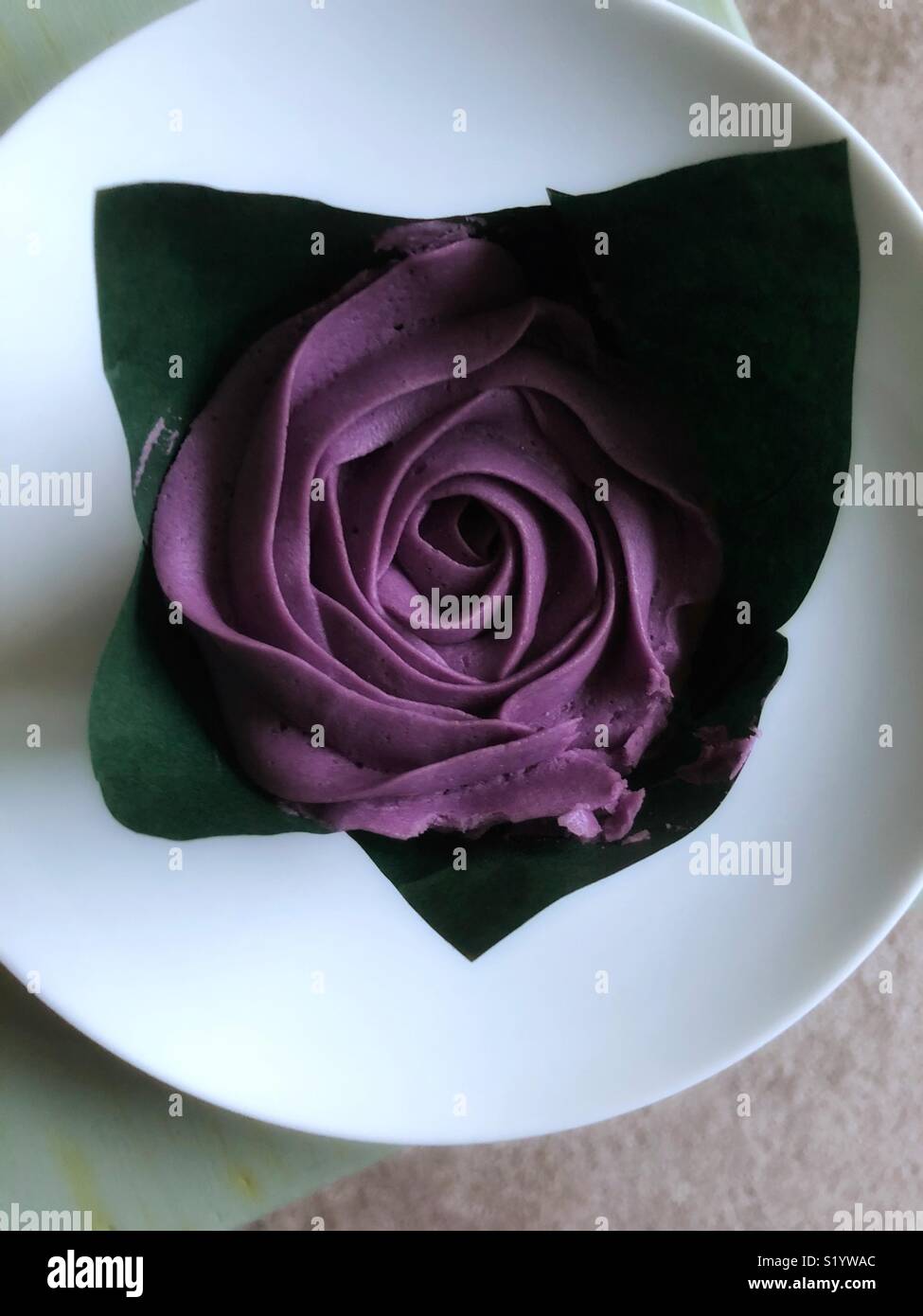 Single piped icing flower on top of a cup cake, wrapped in greaseproof paper to represent leaves sitting on a round white plate. Food art at it’s best. - Smartphone Captured Stock Image