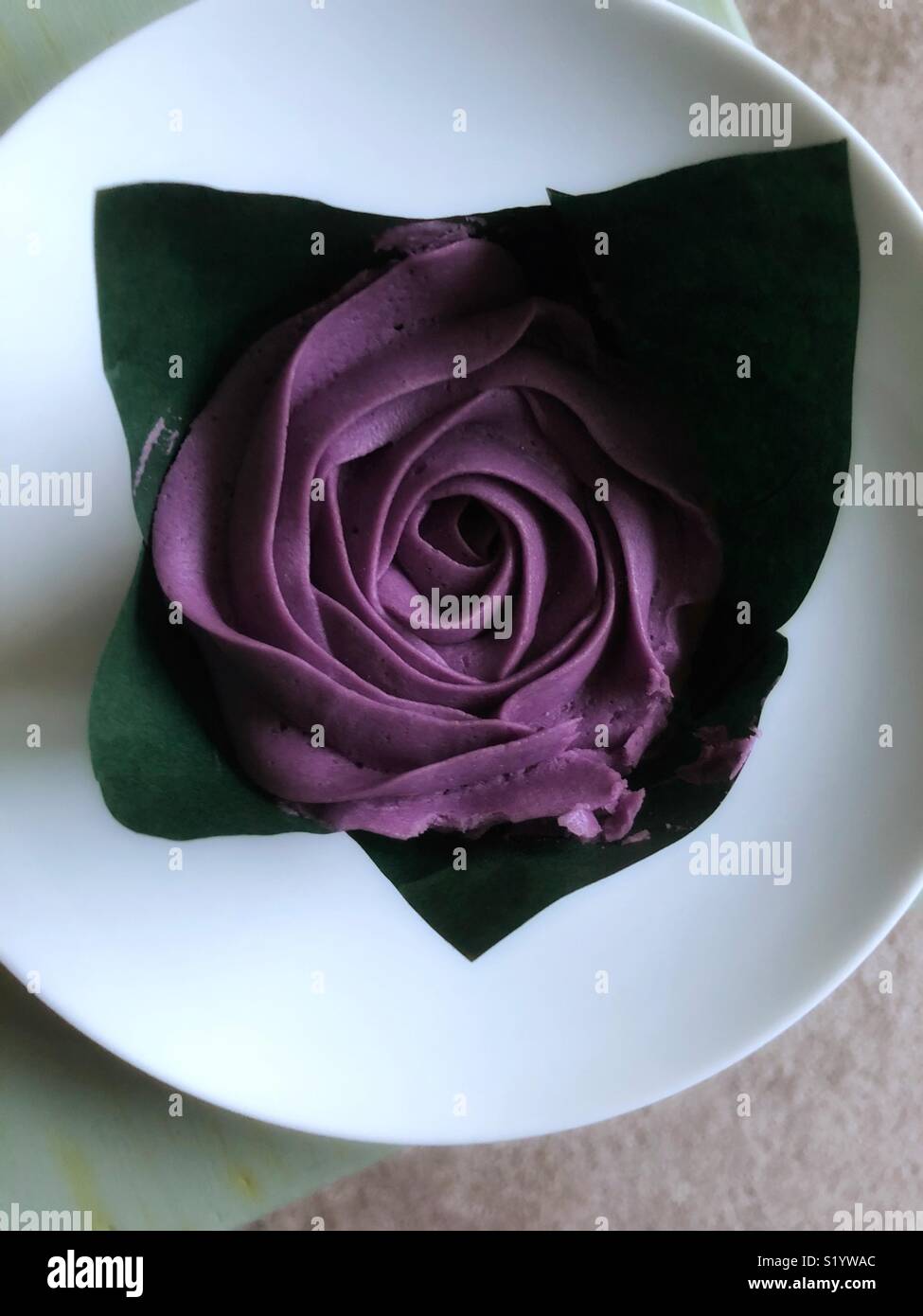 Single piped icing flower on top of a cup cake, wrapped in greaseproof paper to represent leaves sitting on a round white plate. Food art at it’s best. Stock Photo