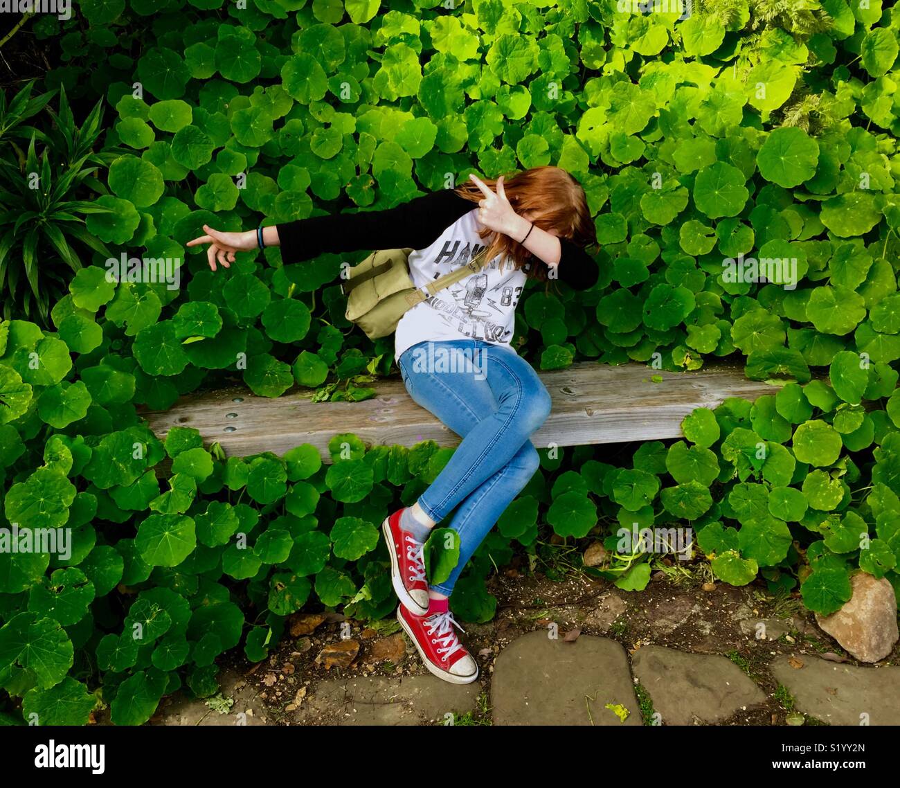 Preteen on a bench wearing red shoes in front of a wall of nasturtiums, hiding her face. - Smartphone Captured Stock Image